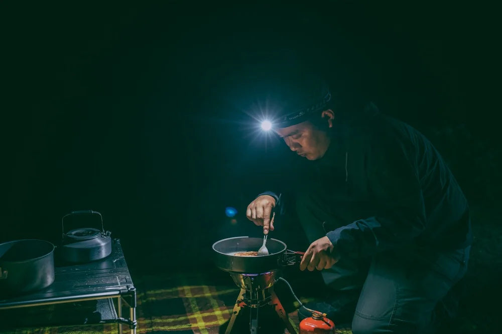 Person cooking outdoors at night using a headlamp and portable stove.