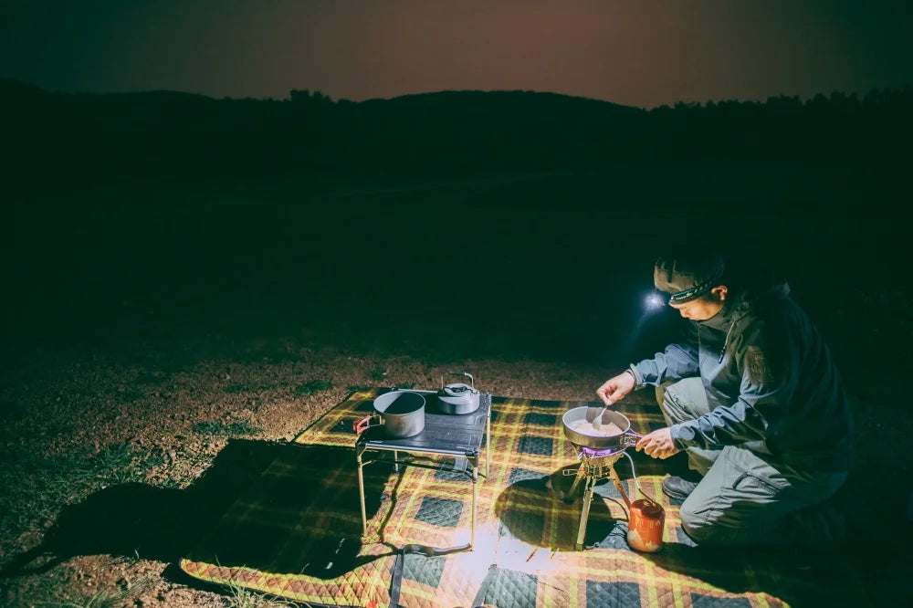 Person camping at night with a lantern, sitting by a small table with a pot and other items.