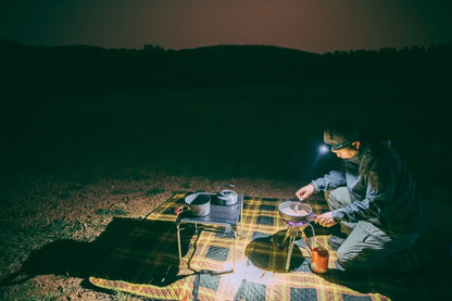 Person camping at night with a lantern, sitting by a small table with a pot and other items.