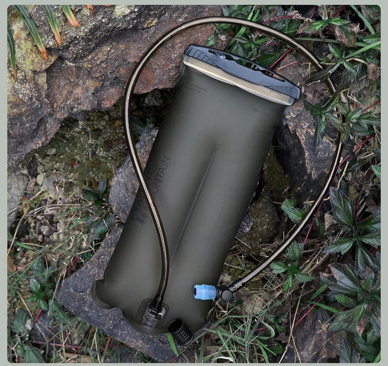 An olive green hydration bladder with a black and gray cap, connected to a black drinking tube, placed on rocks and grass outdoors.