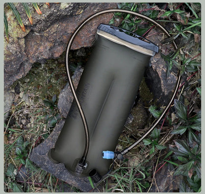 An olive green hydration bladder with a black and gray cap, connected to a black drinking tube, placed on rocks and grass outdoors.