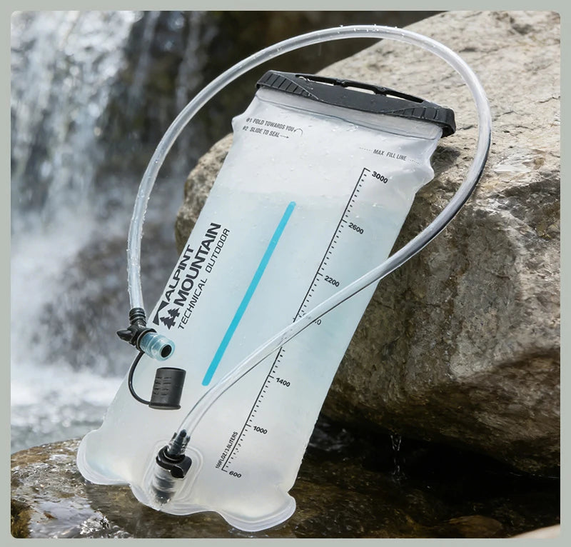 A clear water bladder with a black top and attached clear tube, shown resting on rocks near a waterfall. The bladder has visible printed branding and measurement markings.