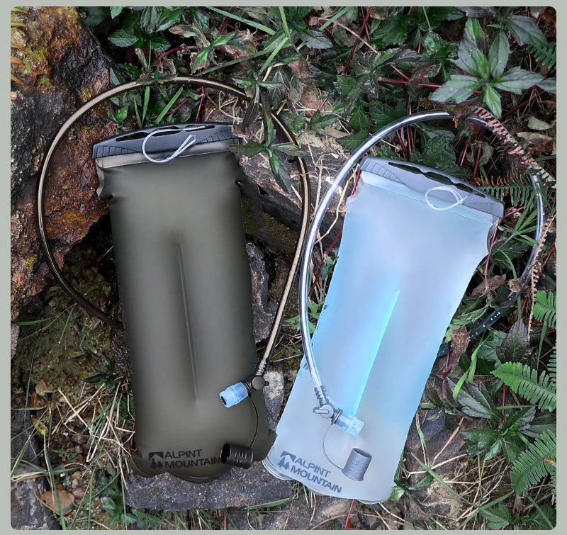 Two Alpine Mountain water bottles on an outdoor trail. One is a matte olive green bottle with a black cap and hose, and the other is a translucent light blue bottle with a clear cap and hose.