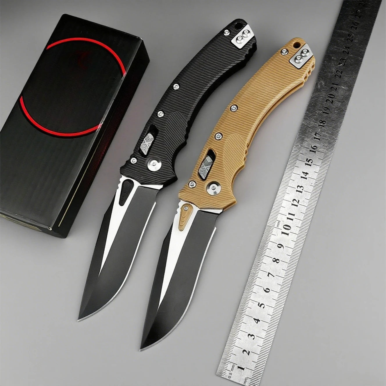 Two folding knives with black and gold handles next to a ruler on a gray background