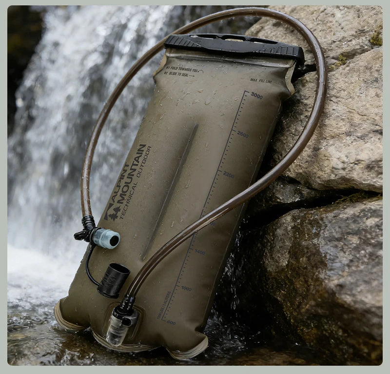An olive green hydration bladder with attached tubing, shown outdoors against rocks and a waterfall. The bladder has visible branding that reads 'MOUNTAIN PEDESTRAL' and measurement markings.