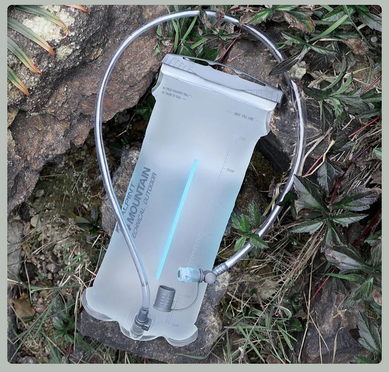 A translucent water bladder with a metal drinking tube loop, placed outdoors on rocks and grass. The bladder has printed branding and measurement markings.