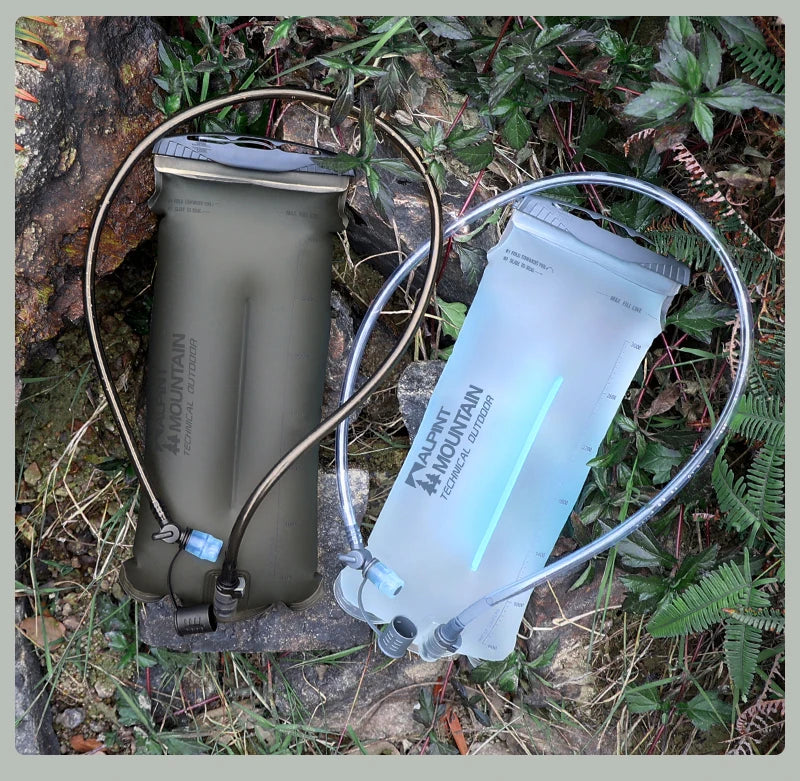 Two Mountain DualFlow hydration packs, one olive green and one translucent light blue, shown outdoors on a rock surrounded by green foliage.
