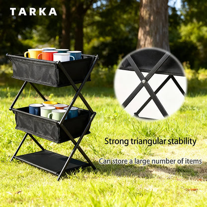 A black metal and fabric outdoor storage cart with two tiers and a lower shelf, shown on grass with trees in the background. The cart holds colorful mugs, and text on the image highlights strong triangular stability and storage capacity.