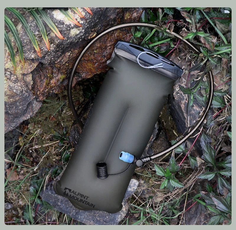 An olive green Alpinestars Mountain Water Pack hydration bladder with a black top closure and a black drinking tube, shown outdoors on rocks and grass.
