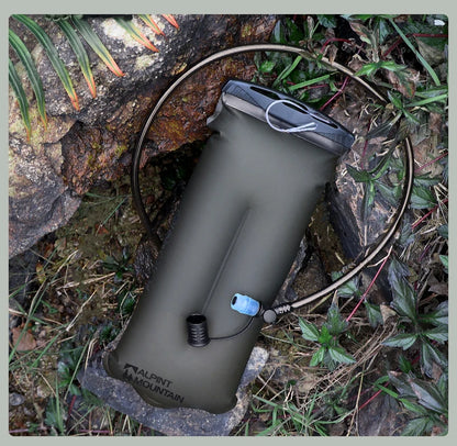 An olive green Alpinestars Mountain Water Pack hydration bladder with a black top closure and a black drinking tube, shown outdoors on rocks and grass.