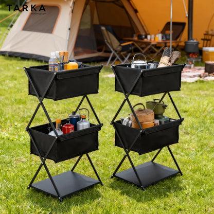 Two black TARKA folding utility carts with storage bins and a lower shelf, shown on a grassy camping ground with a tent and campsite setup in the background.