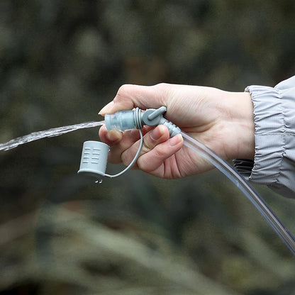 A close-up of a hand holding a gray water filter straw with a clear tube and water flowing out, set against a blurred outdoor background.