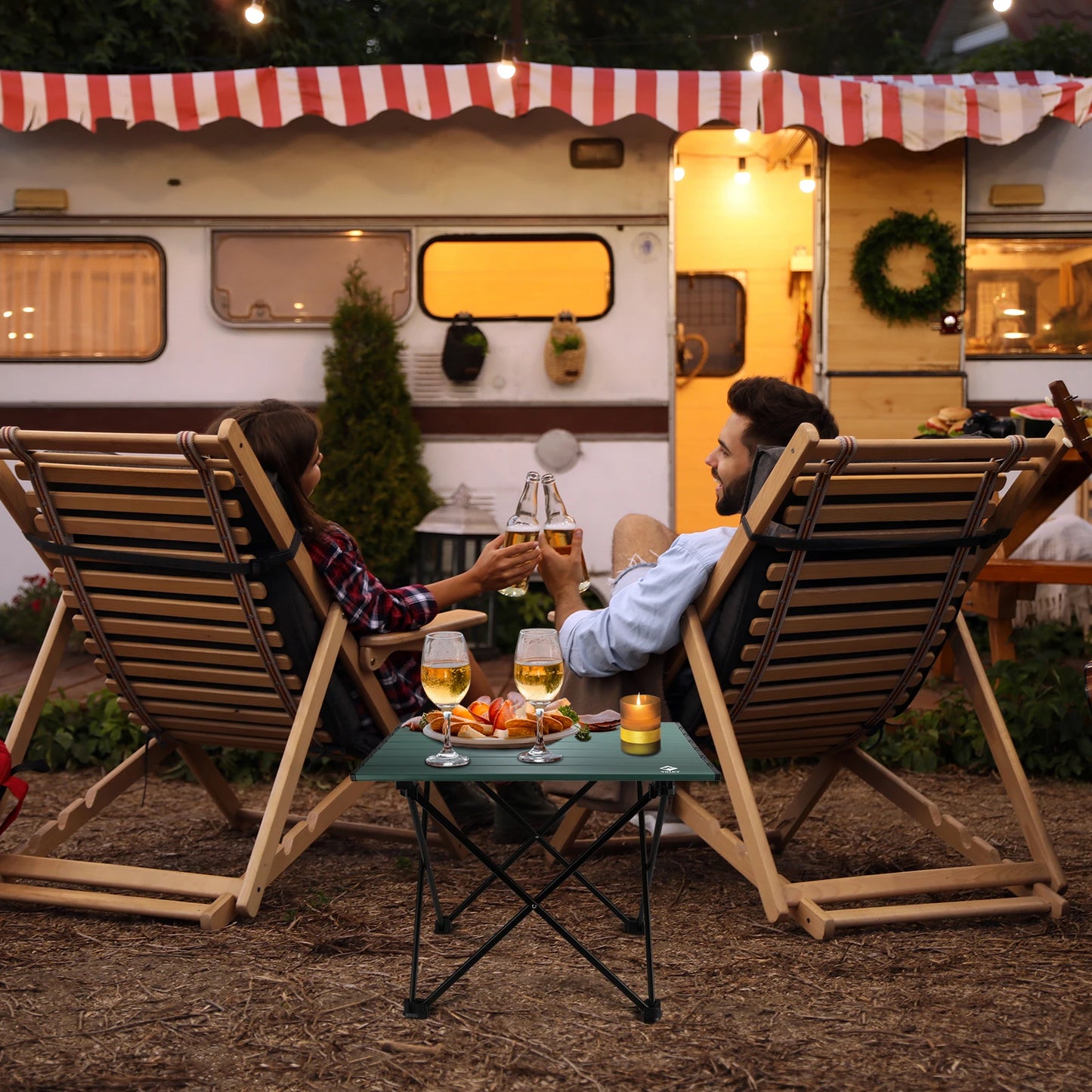 Two people sitting in front of a camper van, enjoying drinks and food on a small table.