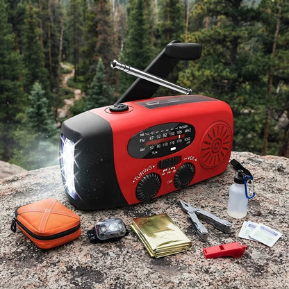 A red and black emergency radio with an extended antenna sits on a rocky forest path, with survival gear placed around it including an orange case, headlamp, folding knife, metal can, and small red tool. Trees and forest are visible in the background.