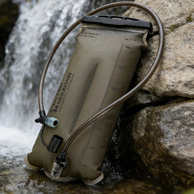 An olive drab green Accu-Mountain hydration bladder with attached drinking tubes, resting against large rocks next to a flowing waterfall.