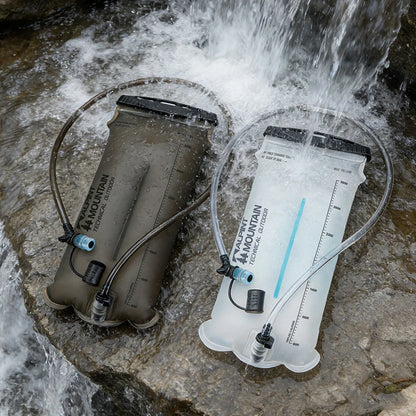Two Mountain Technical Systems hydration reservoirs in a rocky stream with water flowing over them, one in olive drab and one in translucent white.