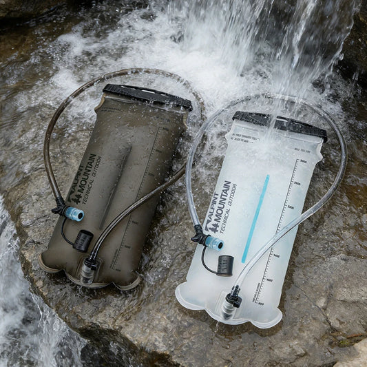 Two Mountain Technical Systems hydration reservoirs in a rocky stream with water flowing over them, one in olive drab and one in translucent white.