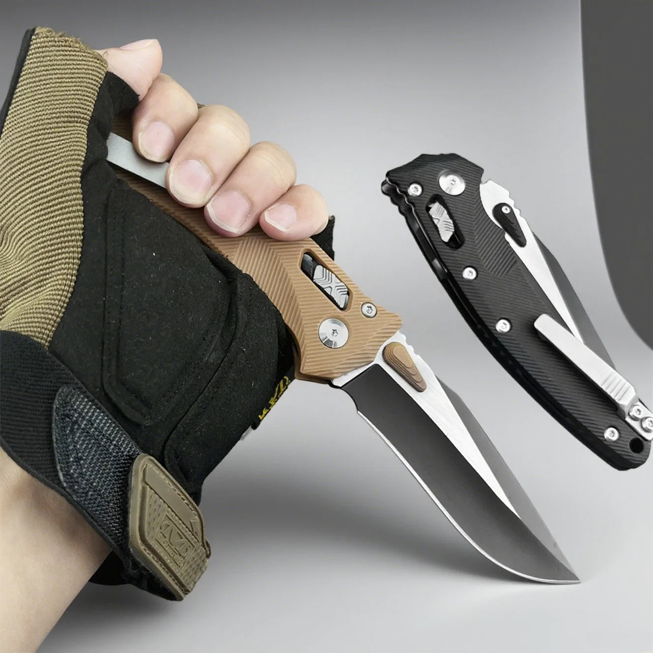 Hand holding a knife with a tactical glove, another knife on a gray background