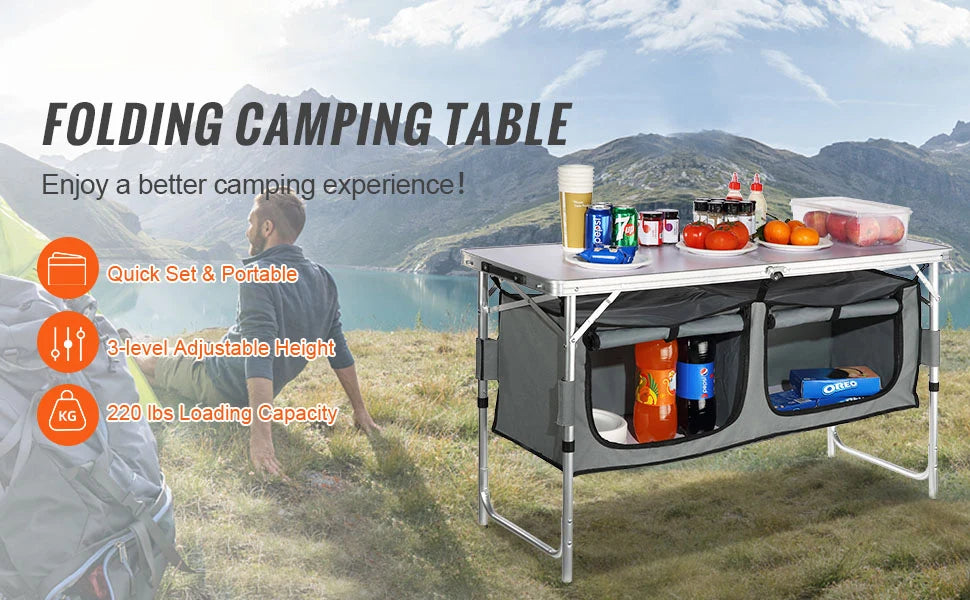 Folding camping table with food and drinks on a grassy field with mountains in the background