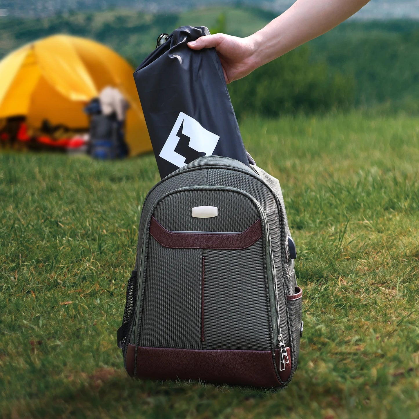 Person holding a black bag over a gray backpack in a grassy outdoor setting with a yellow tent in the background.