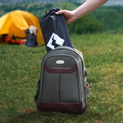 Person holding a black bag over a gray backpack in a grassy outdoor setting with a yellow tent in the background.