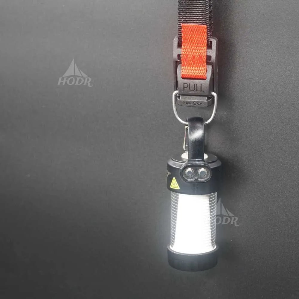 A cylindrical LED lantern light with a black body and bright white illuminated panel, attached to a black strap with an orange pull tab and metal D-ring, shown against a dark gray background.