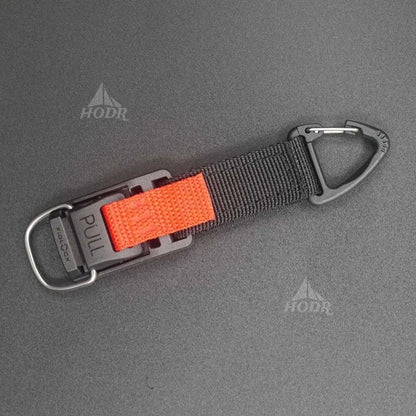 A black webbed strap with a bright orange textured pull tab and gray metal carabiner hardware, shown on a dark gray textured background with HODR watermarks.