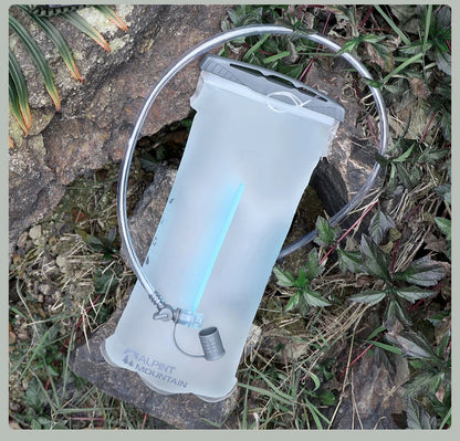 A translucent white Alpinestars Mountain water bottle with a clear drinking tube, resting on a rock outdoors among green plants.