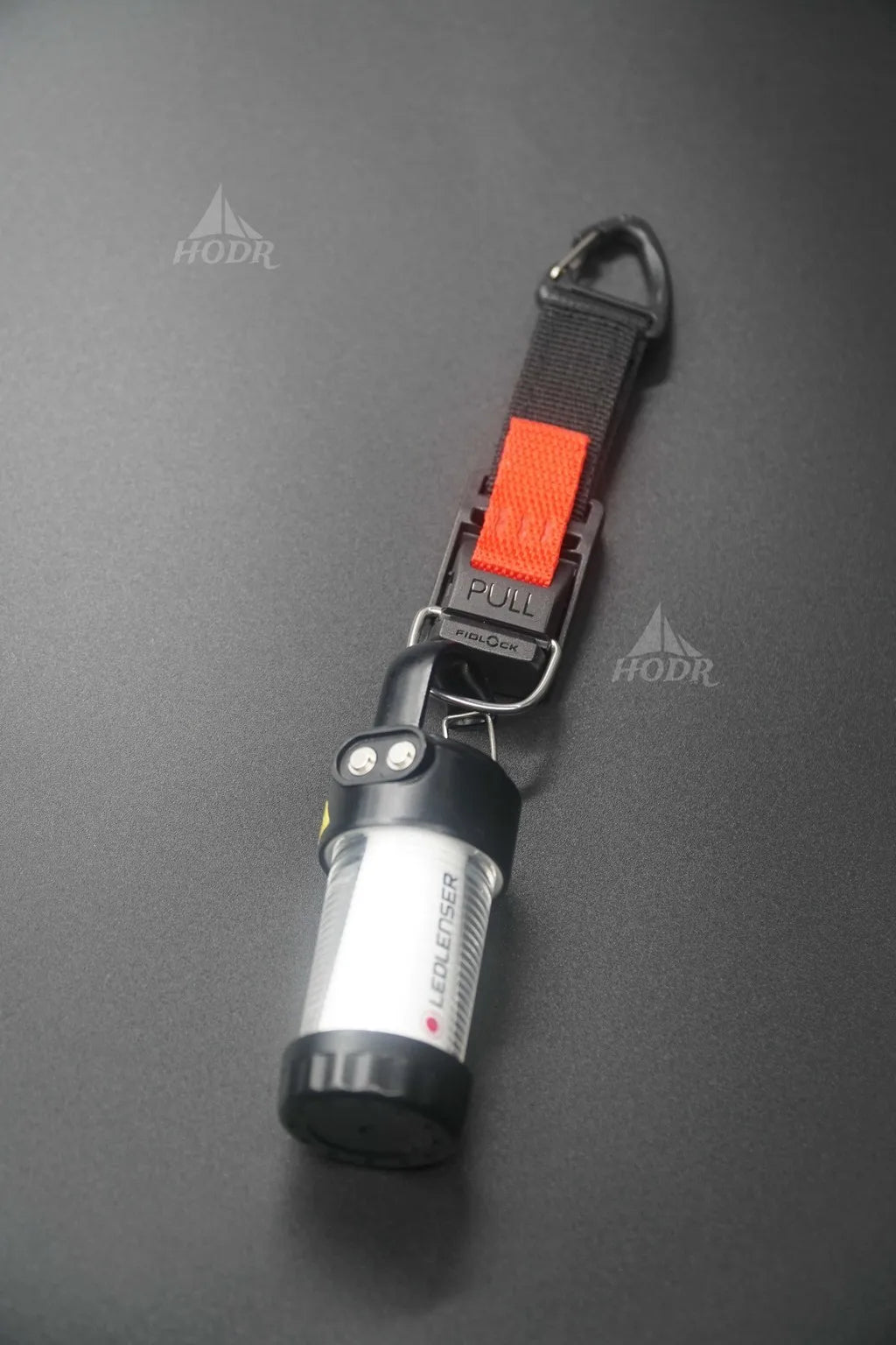 A personal safety keychain featuring a black pepper spray canister with a clear window, attached to a black webbed strap with an orange pull tab and a black carabiner clip, shown on a dark gray background.