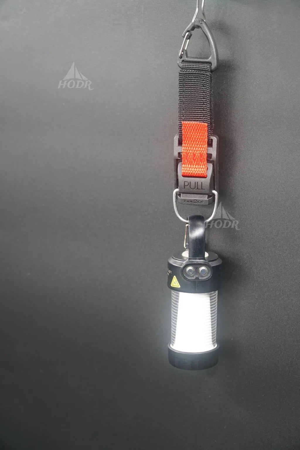 A cylindrical LED lantern light with a black body, hanging from a gray webbed strap with an orange accent and a metal carabiner, shown against a dark gray background.