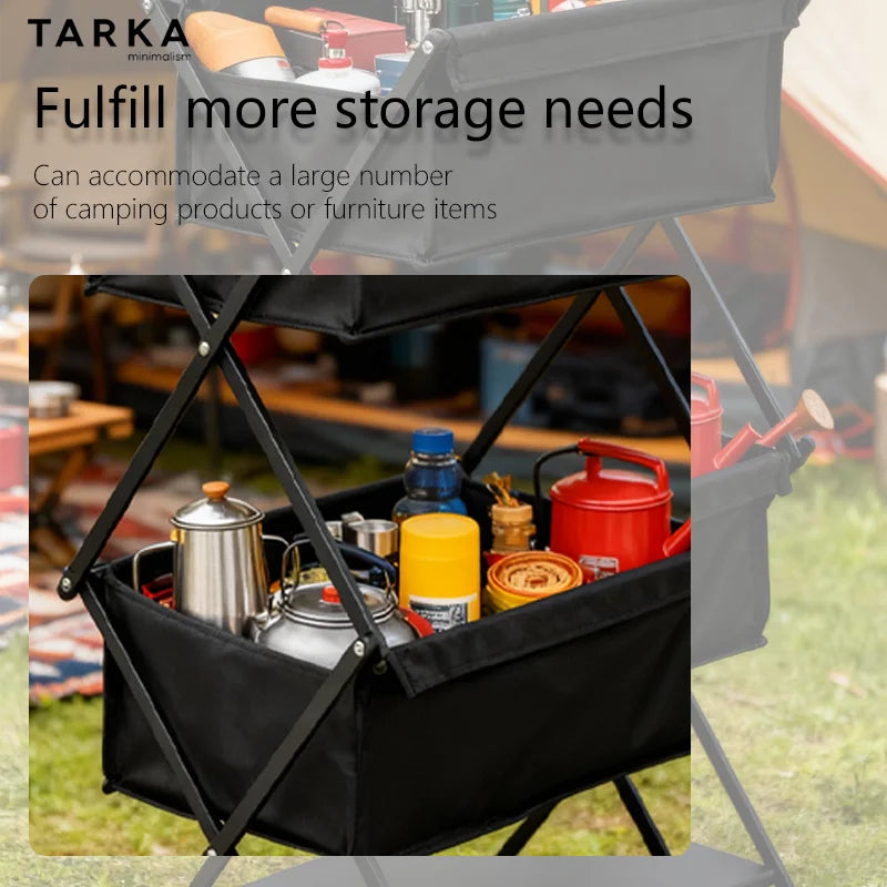 A black folding camp storage cart with fabric bins and metal frame, shown outdoors on grass with camping items inside and a faint branded promotional overlay behind it.