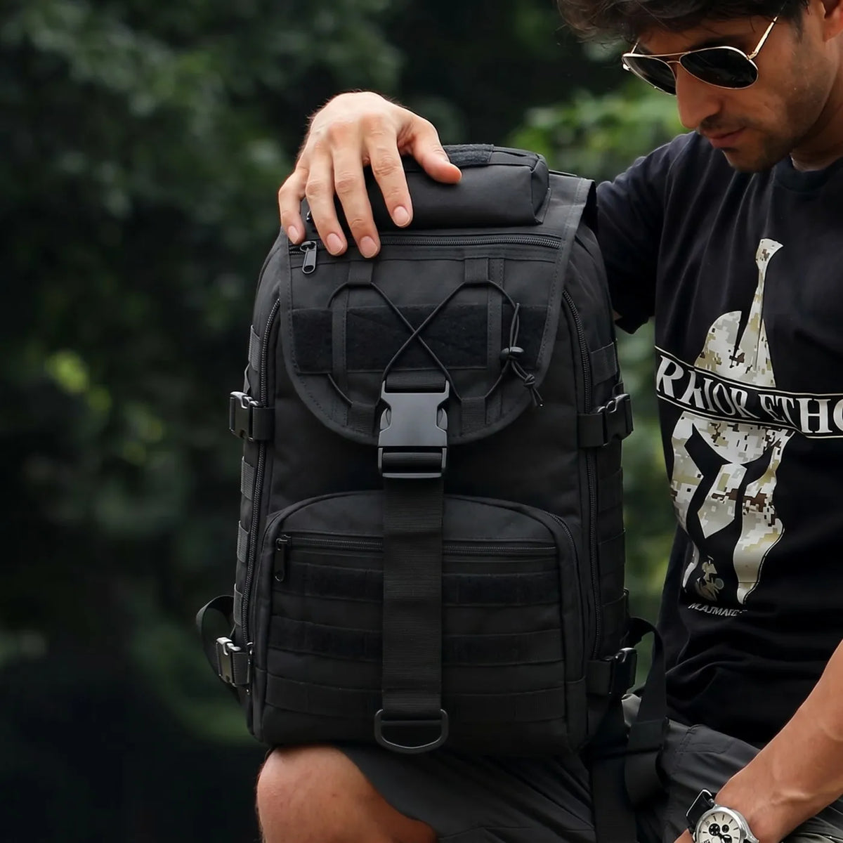 A man wearing a black tactical backpack outdoors. The backpack is black with multiple zippered compartments, webbing, and a front buckle closure.