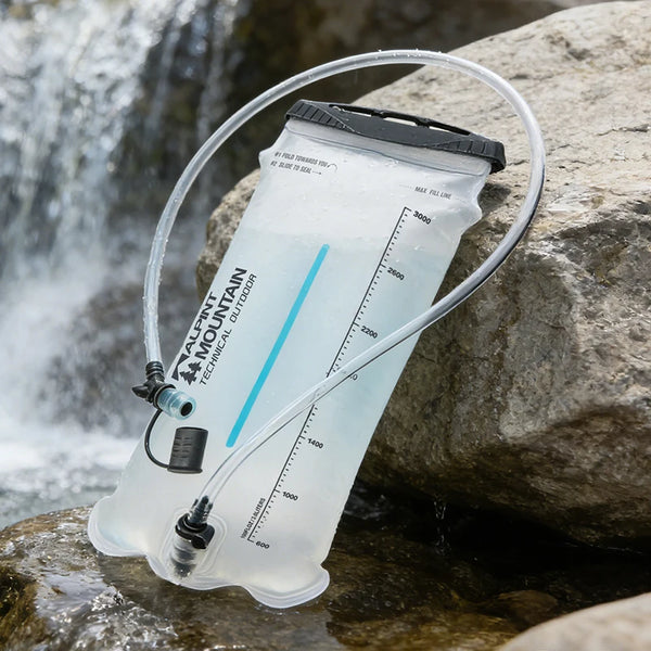 A clear water bladder hydration pack with a black cap and clear tube, resting on wet rocks next to a flowing stream with a blurred waterfall in the background. The brand 'AQUA MOUNTAIN TECHNICAL OUTDOOR' is printed on the bladder.