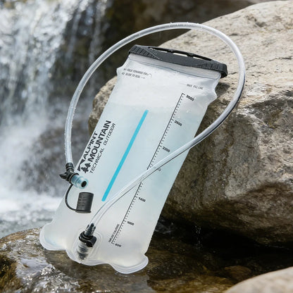 A clear water bladder hydration pack with a black cap and clear tube, resting on wet rocks next to a flowing stream with a blurred waterfall in the background. The brand 'AQUA MOUNTAIN TECHNICAL OUTDOOR' is printed on the bladder.